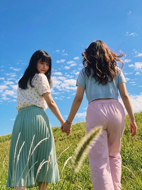 Two girls walking hand in hand up a grassy hill under a blue sky.
